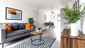 A modern living room with a grey sofa, a coffee table, and a patterned rug. There is a dining area in the background with a table and chairs. - Staycay - Modern 1-bed Apartment in Manchester City Centre (Salford)