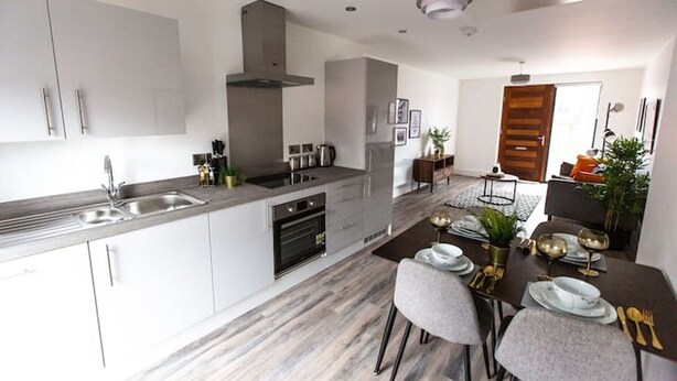 A modern kitchen with white cabinets, stainless steel appliances, and a dining area with a dark table set for two. - Staycay - Modern 1-bed Apartment in Manchester City Centre (Salford)