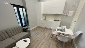 Un appartement moderne avec un canapé gris, une petite table à manger avec des chaises, un coin cuisine et une grande fenêtre.