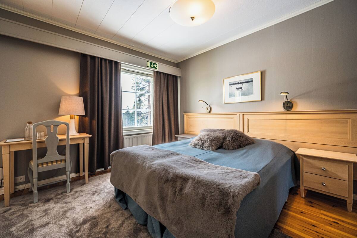 Una camera d'albergo con un letto, una scrivania, una sedia, un comodino e una finestra con tende.