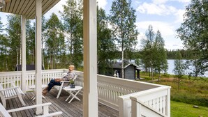 En træterrasse med hvide gelændere, en person der sidder i en stol, og en sø samt skov i baggrunden.