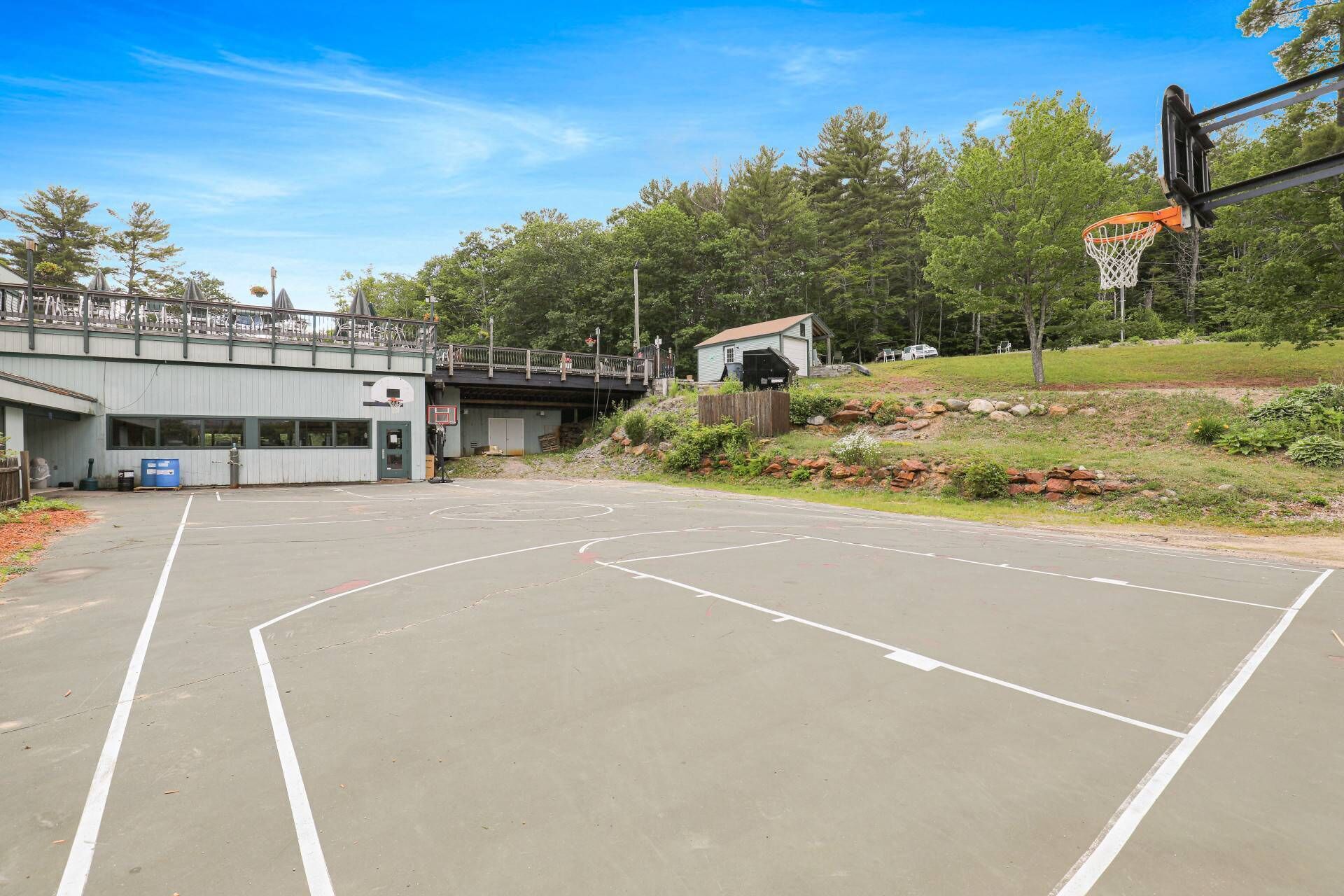 An outdoor basketball court with a wooden bridge overhead, surrounded by trees and a small building.