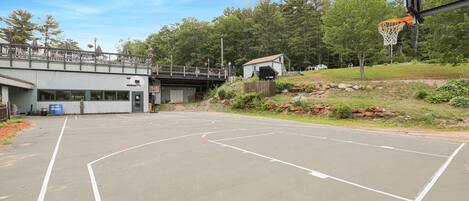 An outdoor basketball court with a wooden bridge overhead, surrounded by trees and a small building.