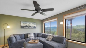 Uma sala de estar moderna com um sofá seccional cinza, uma mesa de centro de madeira e um ventilador de teto. Uma janela grande oferece vista para as montanhas.