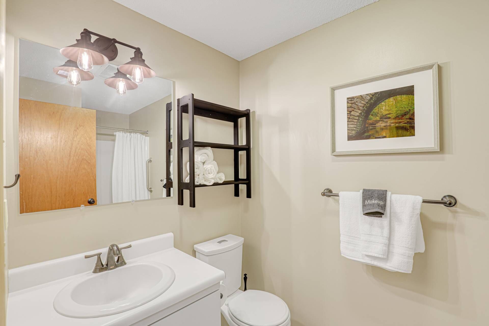 A bathroom with a white sink, a toilet, a towel rack with a towel, a framed picture on the wall, and a mirror with a towel rack underneath.
