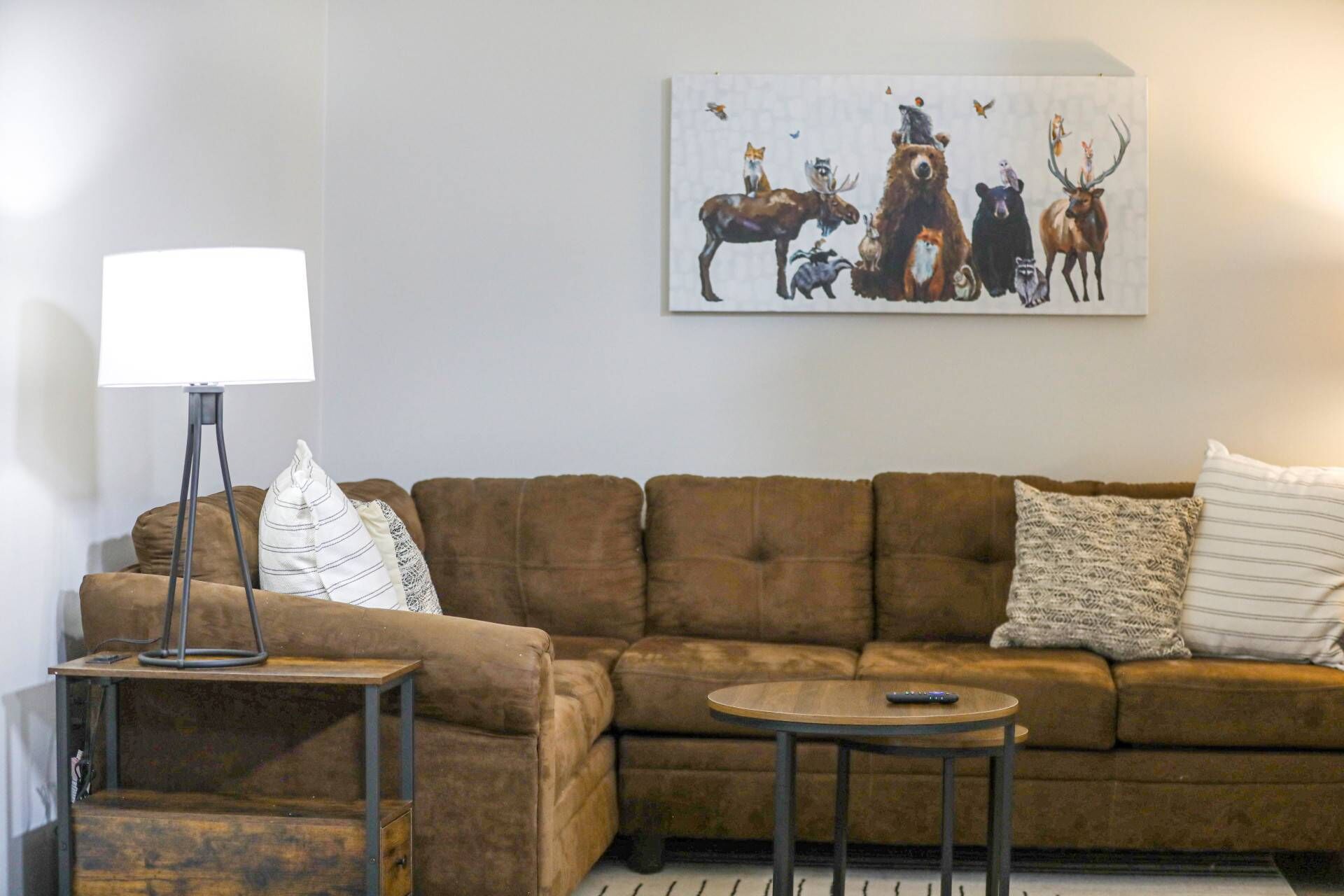 Un salon avec un canapé marron, une petite table d’appoint, une lampe et un tableau représentant des animaux sauvages accroché au mur.