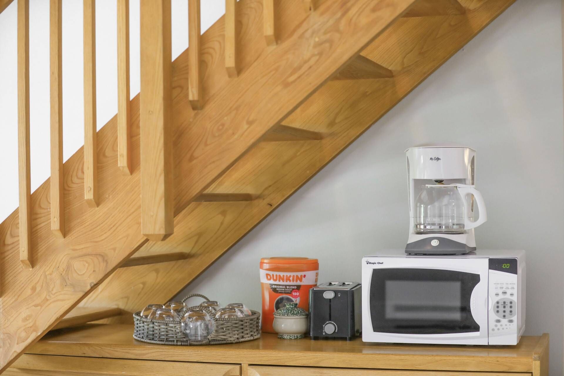 Un comptoir de cuisine avec une machine à café, un four à micro-ondes, un grille-pain, un panier rempli de verres et une boîte de café Dunkin'.