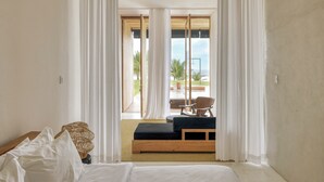A bedroom with a bed, a wooden bench, a chair, and a view of the ocean through sheer curtains.