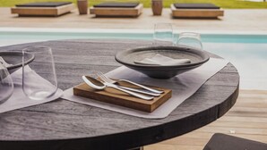 A dining table with a bowl, cutlery, and glasses set on a wooden deck overlooking a pool and beach.