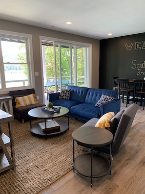 A modern living room with a blue sofa, two round coffee tables, and a rug. There are large windows with a view of greenery outside. - Large Lakefront home w/ Private Dock in Big Creek, beautiful views! (LaFollette)