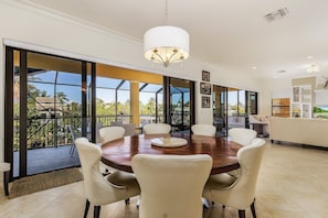 A spacious dining area with a round table, white chairs, and a chandelier. Large windows offer a view of the outdoors.