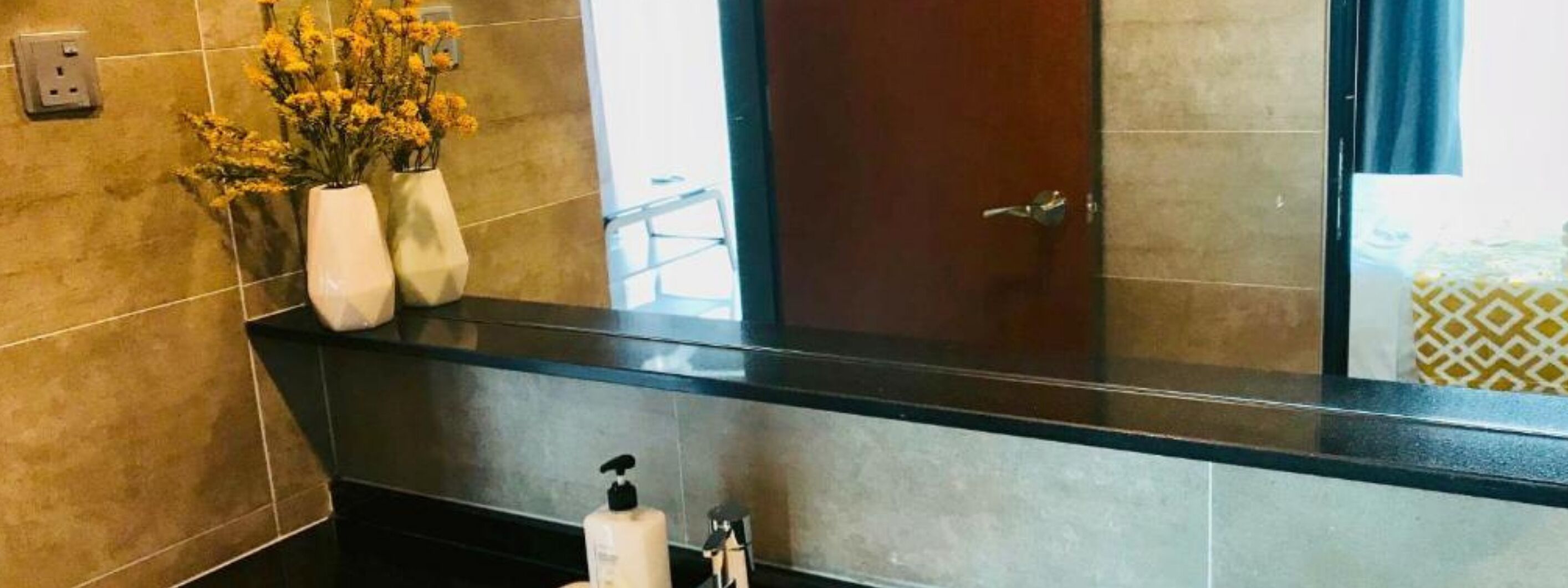 A bathroom with a black countertop, a mirror, and a door. There are two white vases with yellow flowers on the countertop.