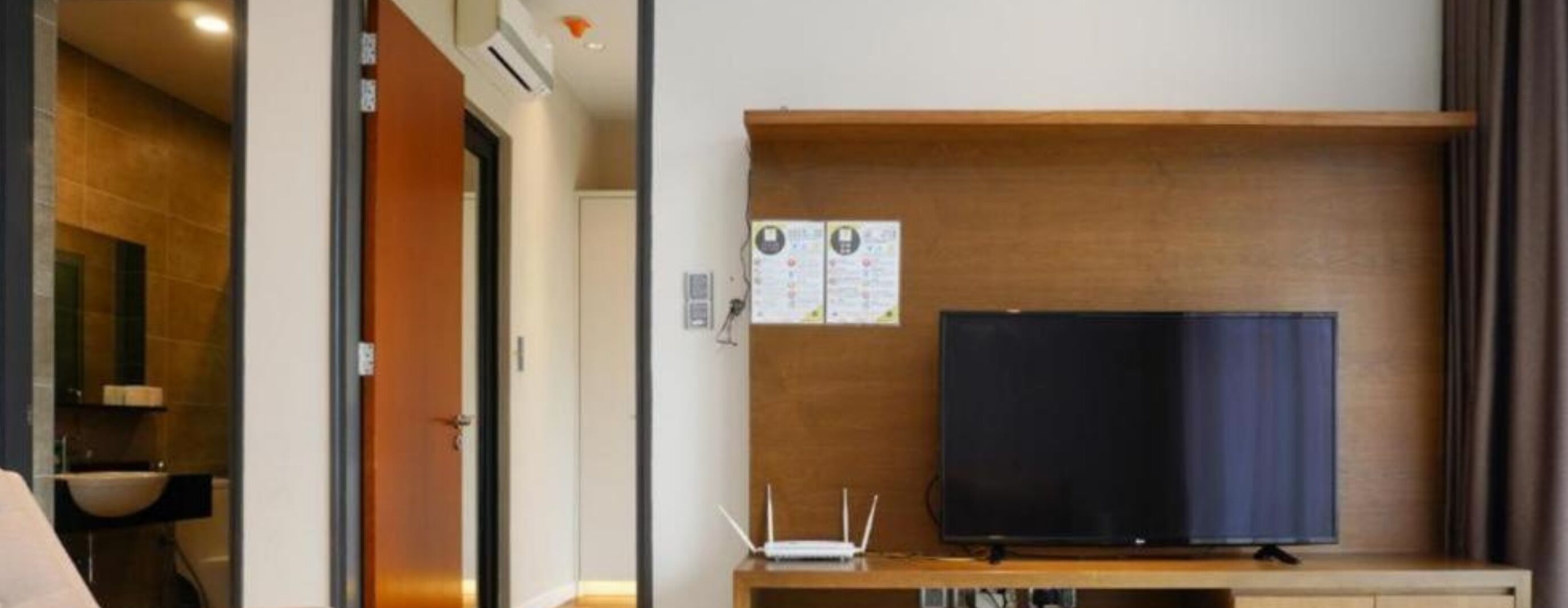 Habitación de hotel con estante de madera, televisor de pantalla plana, una pizarra con anuncios y una pequeña mesa con un router.