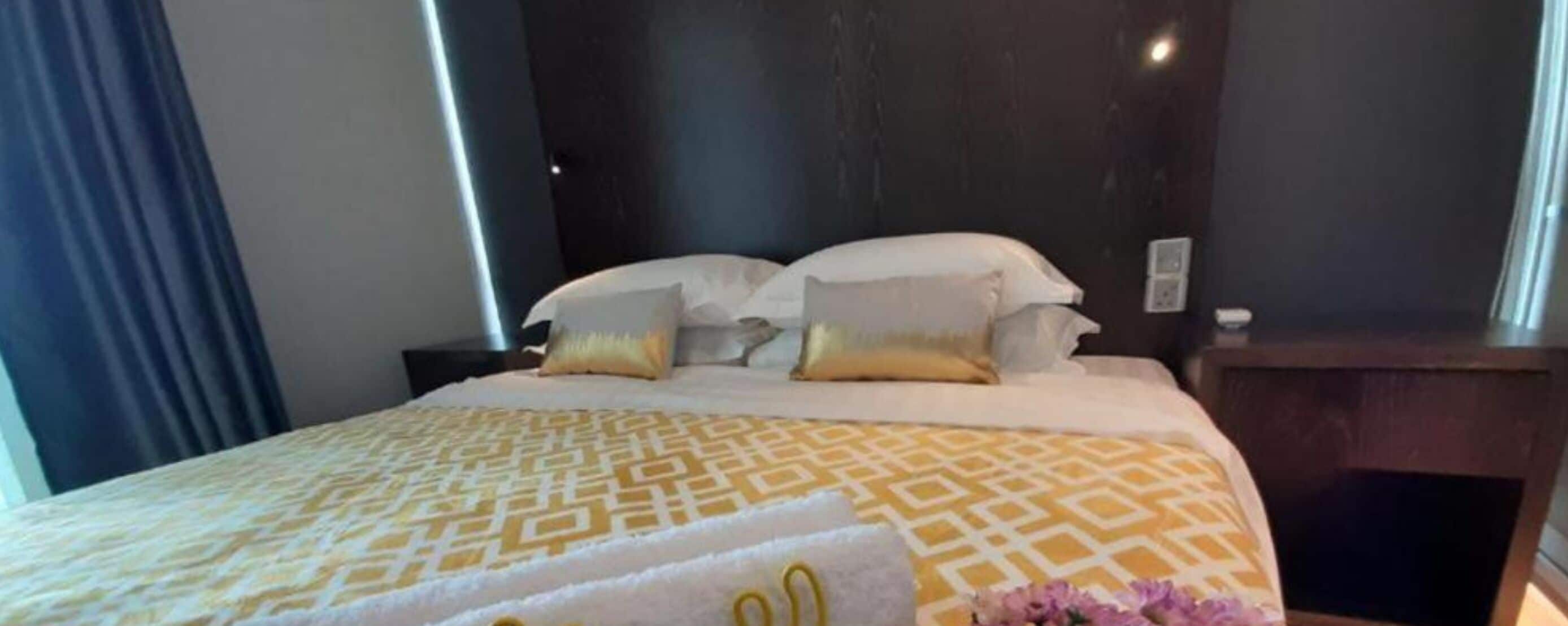 Una cama bien hecha con colcha y almohadas estampadas, cabecera oscura y mesita de noche con lámpara.