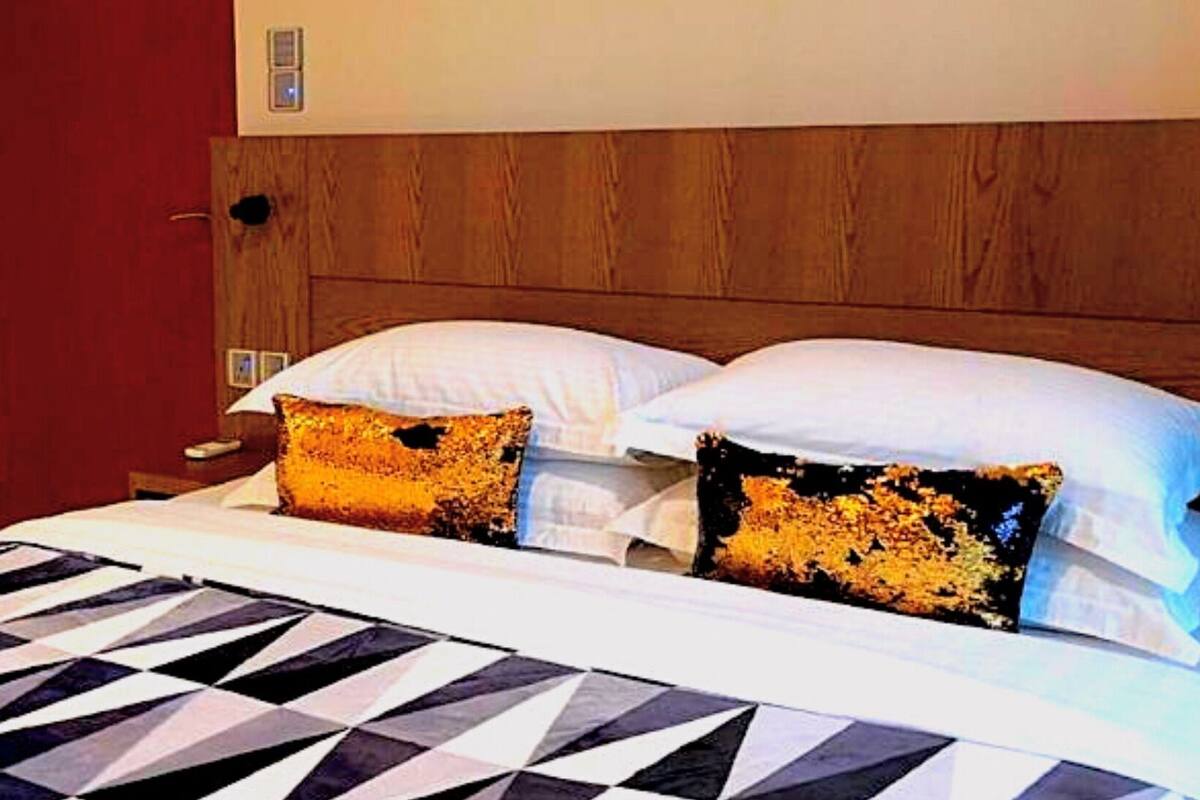 Une chambre d’hôtel avec une tête de lit en bois, une couvre-lit à motifs noir et blanc et deux oreillers ornés d’un motif doré et noir.