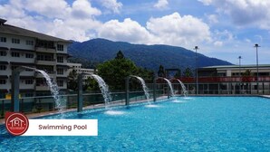 A swimming pool with water fountains, surrounded by buildings and mountains in the background.