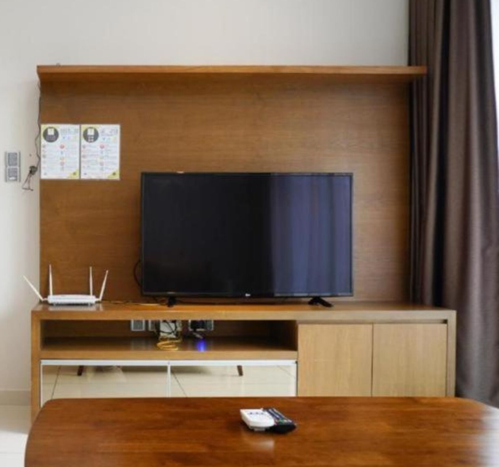 A room with a wooden TV stand, a flat-screen TV, a small table with a remote control, and a wall-mounted shelf with papers.