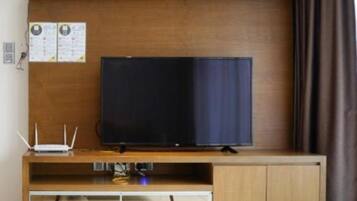 A room with a wooden TV stand, a flat-screen TV, a small table with a remote control, and a wall-mounted shelf with papers.