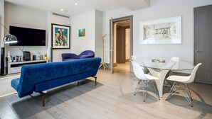 A modern living room with a blue sofa, a glass dining table with white chairs, a flat-screen TV, and artwork on the walls.