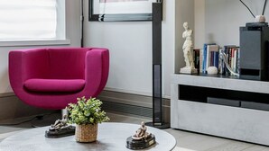 A modern living room with a round coffee table, a vibrant pink armchair, a mounted TV, and a bookshelf with books and decorative items.