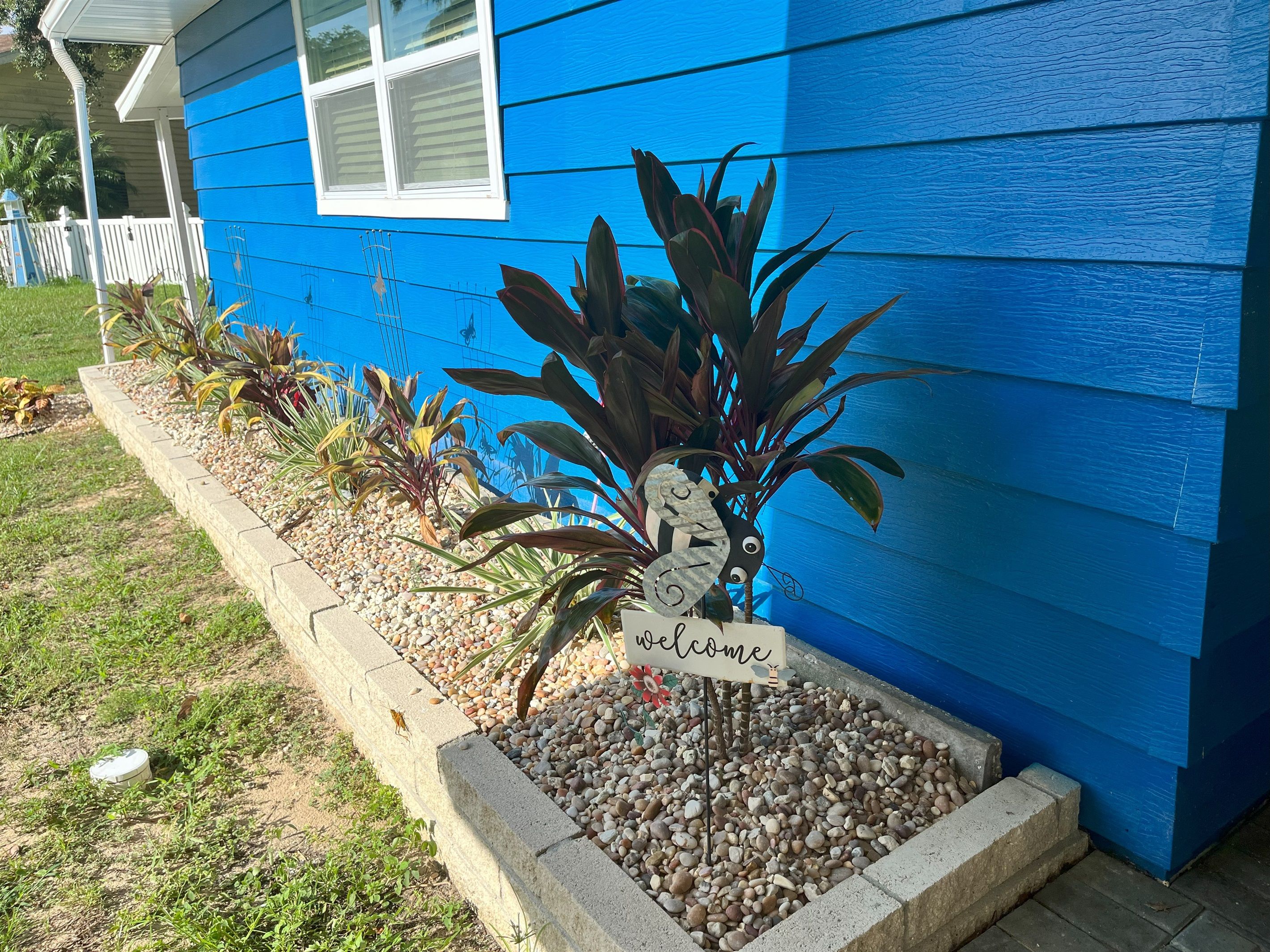 Une maison avec un revêtement extérieur bleu, un panneau d’accueil et un parterre agrémenté de galets et de plantes.