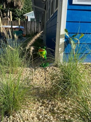 A garden area with a frog statue, a wooden fence, and a blue building. - River Beach Retreat: Tiki Bar, Hot Tub, Kayaks&BBQ (Spring Hill)