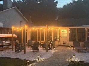 A covered outdoor seating area with chairs and tables, string lights, and a building in the background. - River Beach Retreat: Tiki Bar, Hot Tub, Kayaks&BBQ (Spring Hill)