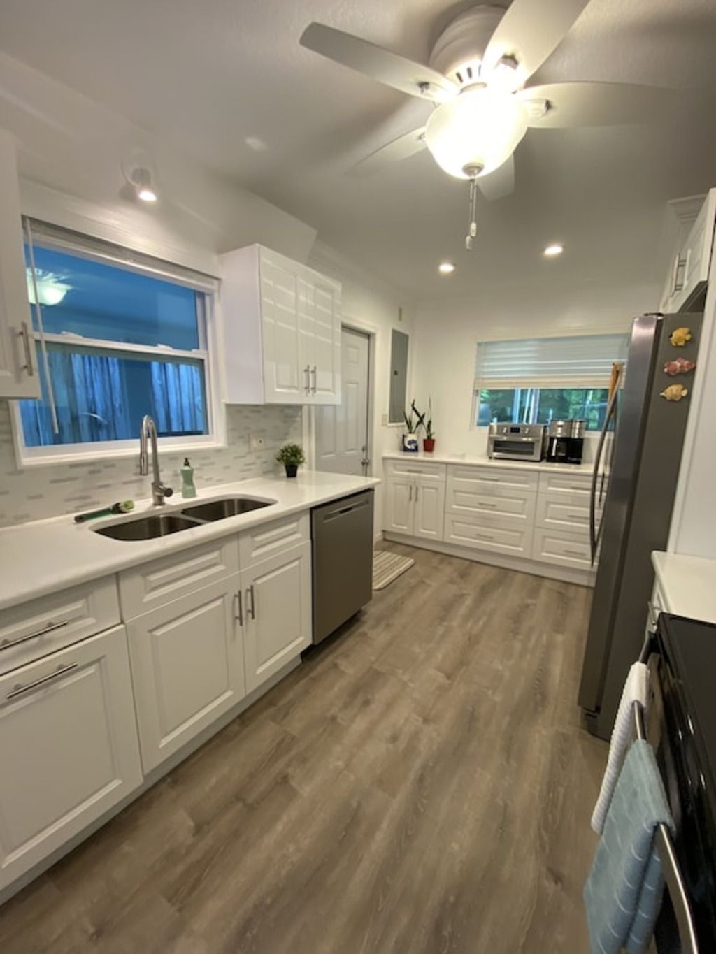 Una cocina moderna con armarios blancos, lavavajillas, ventilador de techo y una ventana con cortinas.