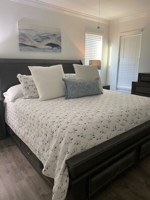 A bedroom with a bed, a nightstand, a dresser, a painting on the wall, and a window with blinds.