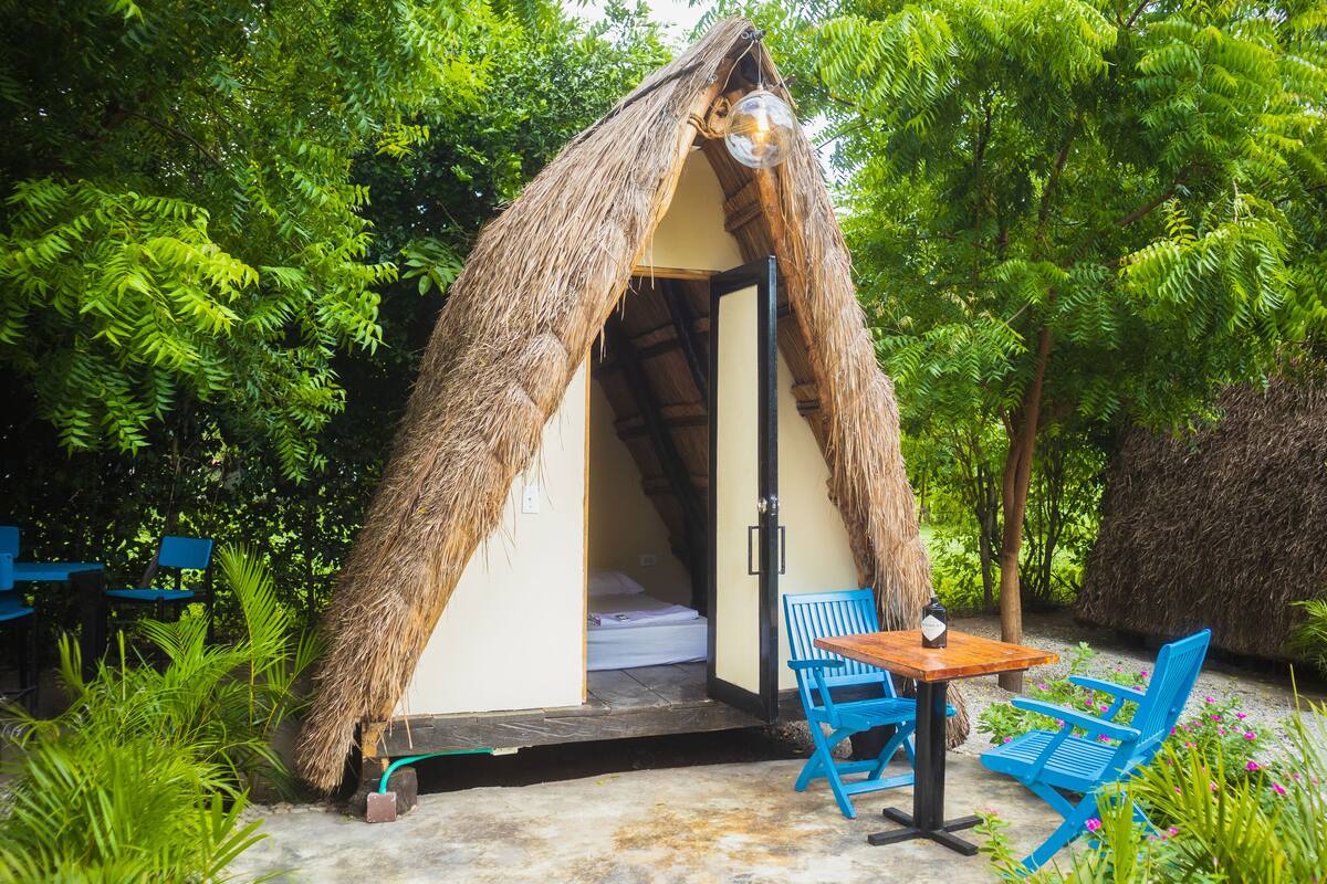A thatched-roof hut with a bed inside, a table, and blue chairs outside.