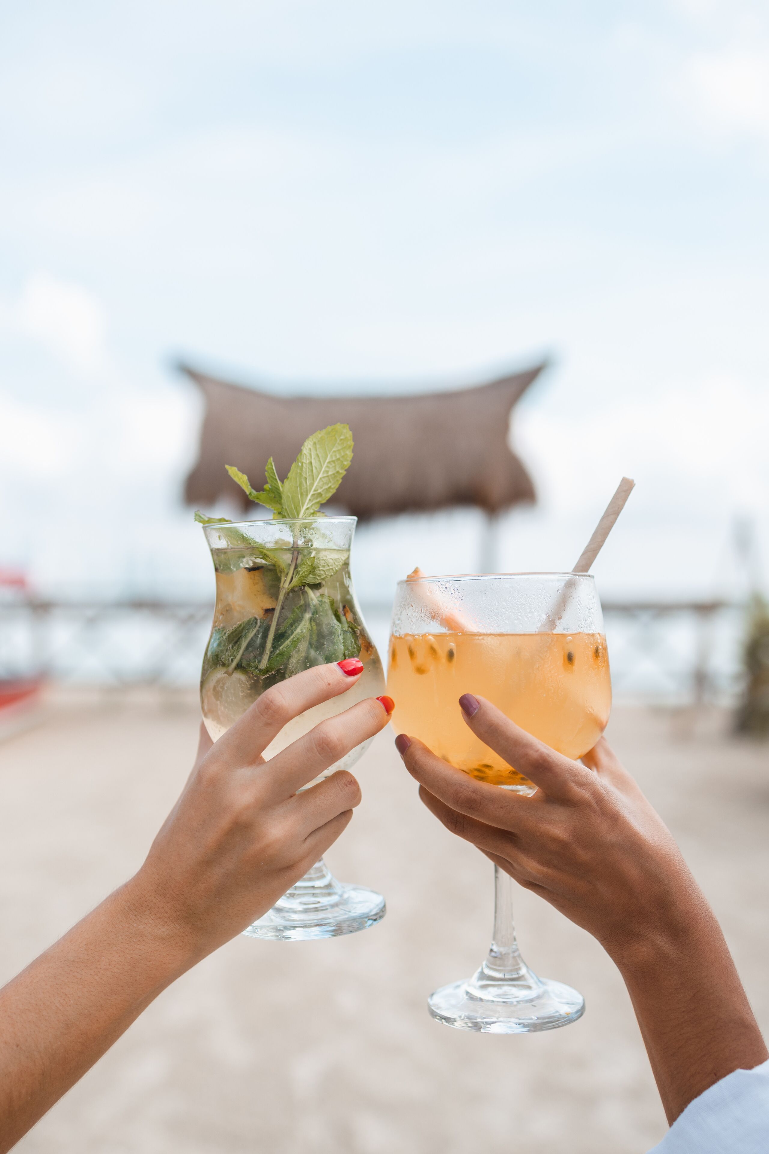 Duas pessoas segurando copos com bebidas em uma praia, com uma estrutura ao fundo.