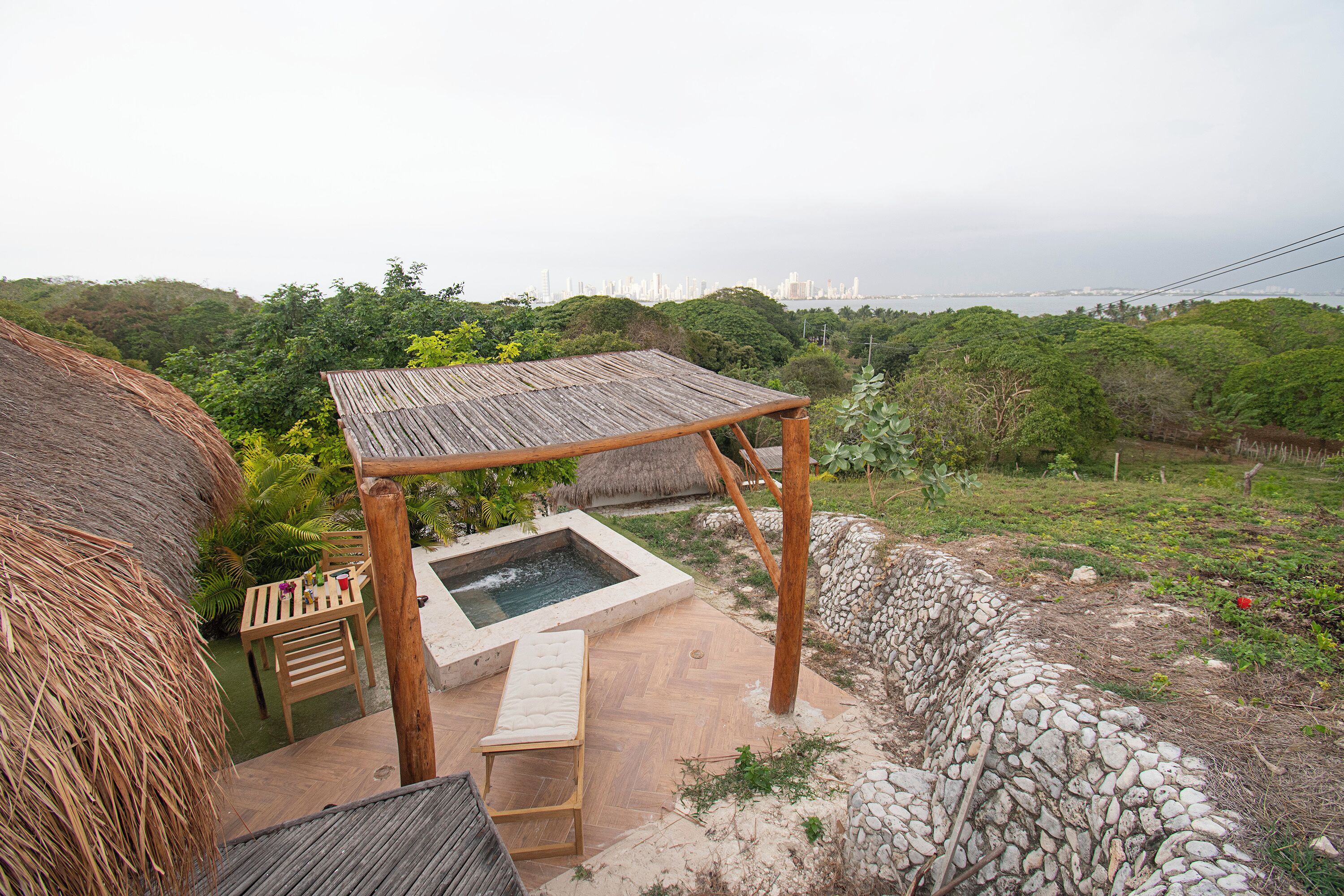 Una pérgola de madera con jacuzzi y zona de estar, con vistas a un jardín exuberante y al horizonte de la ciudad.