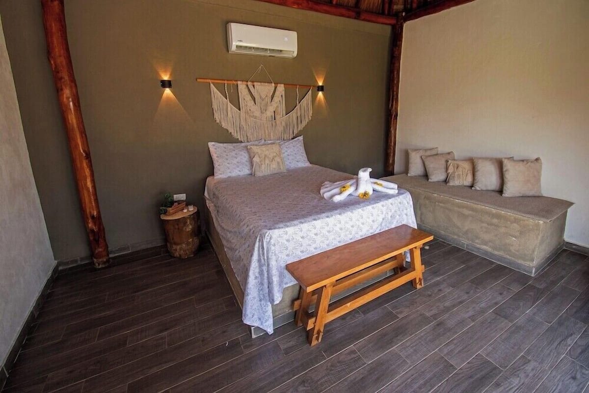 A bedroom with a bed, bedside table, bench, wall-mounted air conditioner, and a bench with cushions.