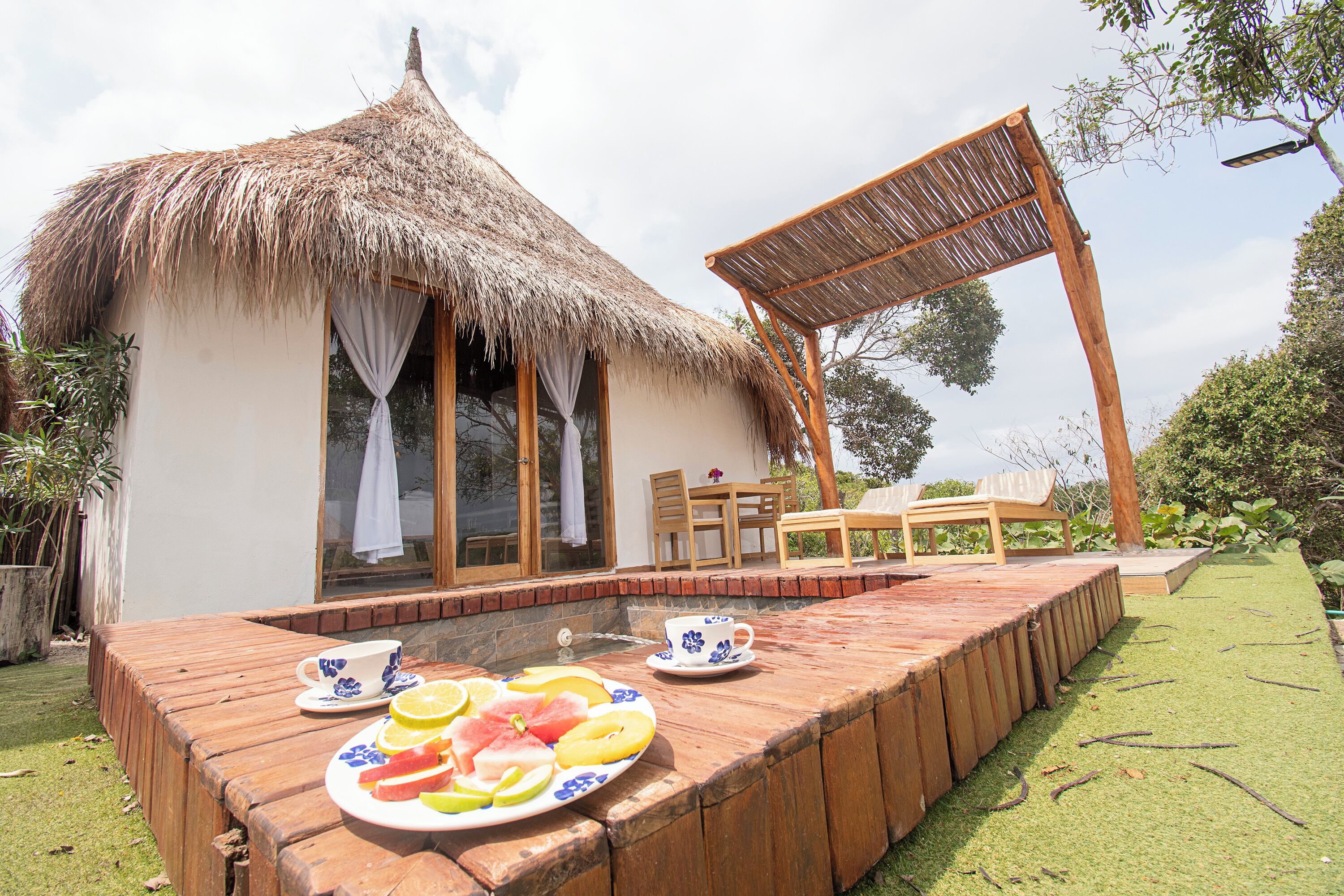Una cabaña con techo de paja y una terraza de madera, una mesa con un plato de fruta y una silla.