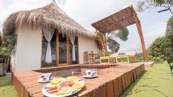 Una cabaña con techo de paja y una terraza de madera, una mesa con un plato de fruta y una silla.
