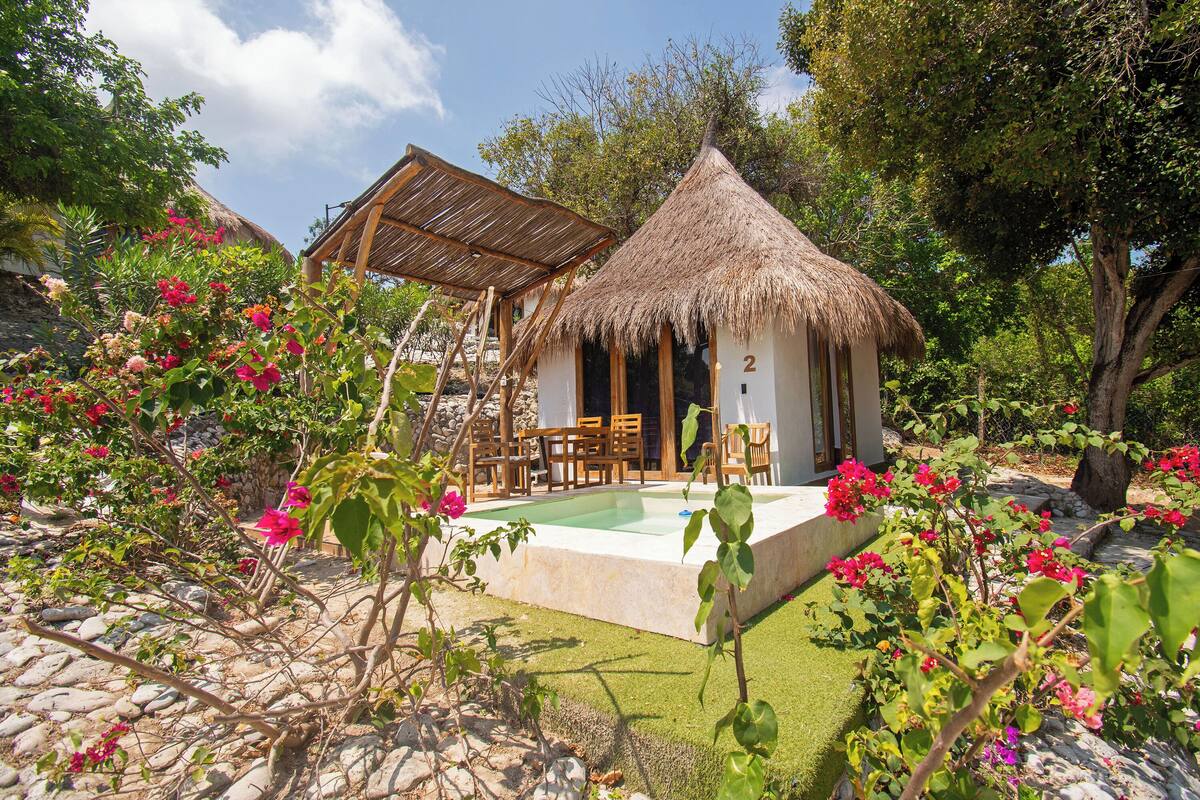 A thatched-roof structure with a pool, surrounded by lush greenery and flowering plants.