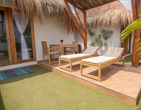 A sunlit outdoor seating area with two wooden lounge chairs, a wooden table, and chairs, surrounded by a thatched roof structure. - AMALIFE Beach Club & Hotel (Cartagena)