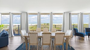Un ampio soggiorno con un grande tavolo da pranzo e sedie, un divano e vista sulla spiaggia attraverso ampie finestre.