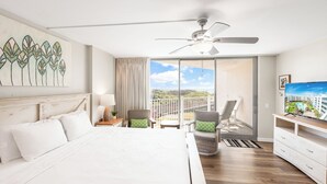 A hotel room with a large bed, a ceiling fan, a balcony with chairs, and a TV. - Sandcastle Condominium Unit 102 (Port Aransas)
