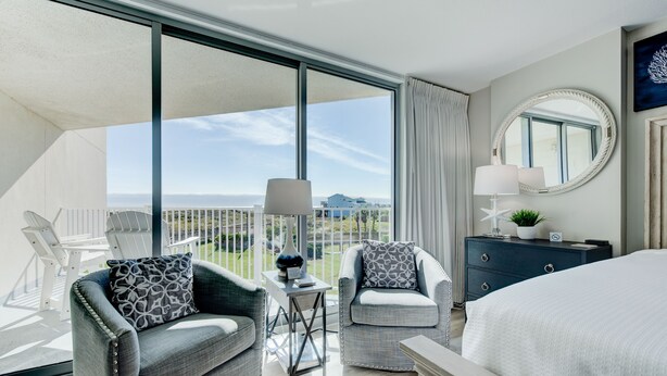 A modern hotel room with a balcony, two armchairs, a bed, a dresser, and a large window offering a city view. - Sandcastle Condominiums Unit 107 (Port Aransas)