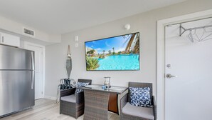 A modern kitchen with a stainless steel refrigerator, a dining area with wicker chairs and a table, and a large TV screen displaying a pool scene. - Sandcastle Condominiums Unit 107 (Port Aransas)