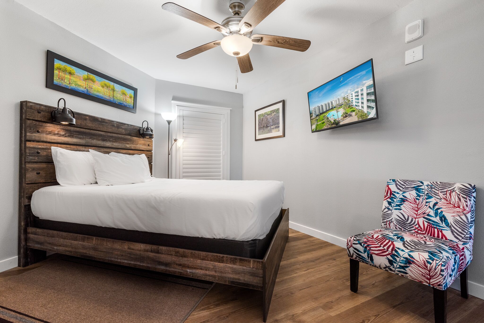 A bedroom with a wooden bed, a ceiling fan, a chair with a tropical print, and two framed pictures on the wall.