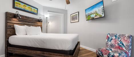 A bedroom with a wooden bed, a ceiling fan, a chair with a tropical print, and two framed pictures on the wall.