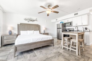 Quarto com cama, mesas de cabeceira, ventilador de teto e cozinha compacta com geladeira, fogão e banquetas.