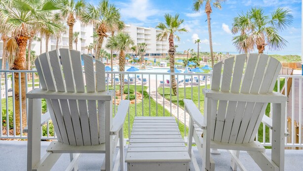 Due poltrone Adirondack bianche su un balcone con vista su una piscina, palme e l'edificio di un hotel sullo sfondo.