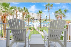 Two white Adirondack chairs on a balcony overlooking a pool area with palm trees and a hotel building in the background. - Sandcastle Condominiums Unit 126 (Port Aransas)