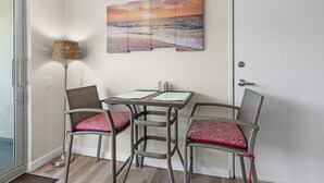 A room with a dining table, two wicker chairs, a wall-mounted lamp, and a sunset painting. - Sandcastle Condominiums Unit 135 (Port Aransas)