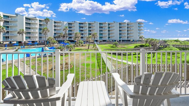 A balcony with white Adirondack chairs and a view of a swimming pool, a grassy area, and a multi-story building with balconies. - Sandcastle Condominiums Unit 135 (Port Aransas)