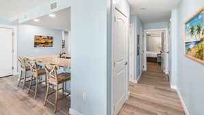 A modern kitchen with a marble countertop and wooden chairs, a hallway with light blue walls and wooden flooring, and a door leading to another room. - Sandcastle Condominiums Unit 212 (Port Aransas)