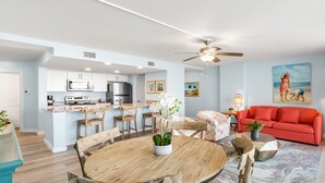 A modern kitchen with white cabinets, a dining area with a wooden table and chairs, a red sofa, and a ceiling fan. - Sandcastle Condominiums Unit 212 (Port Aransas)